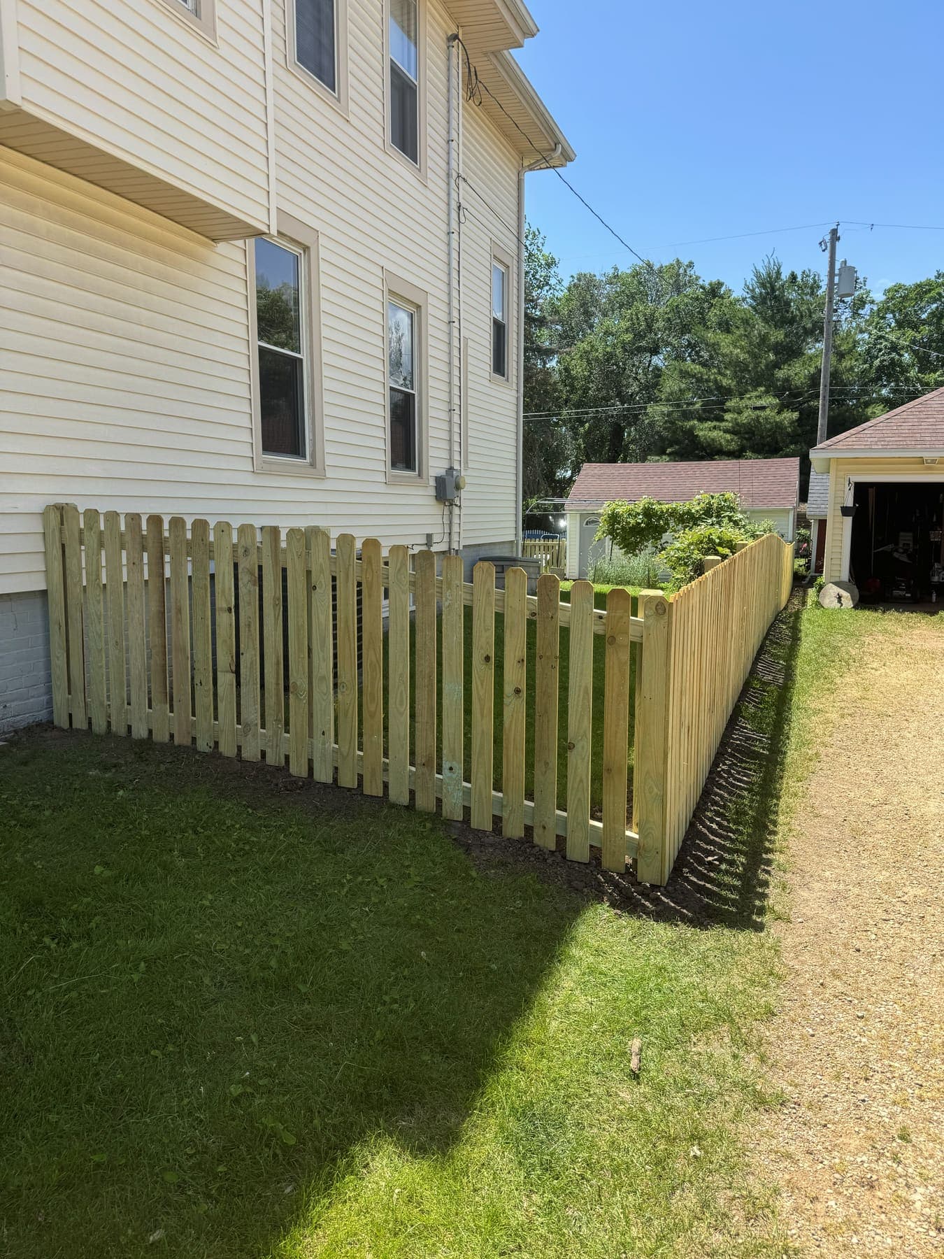 Treated wood picket fence with a scalloped top edge running alongside a house
