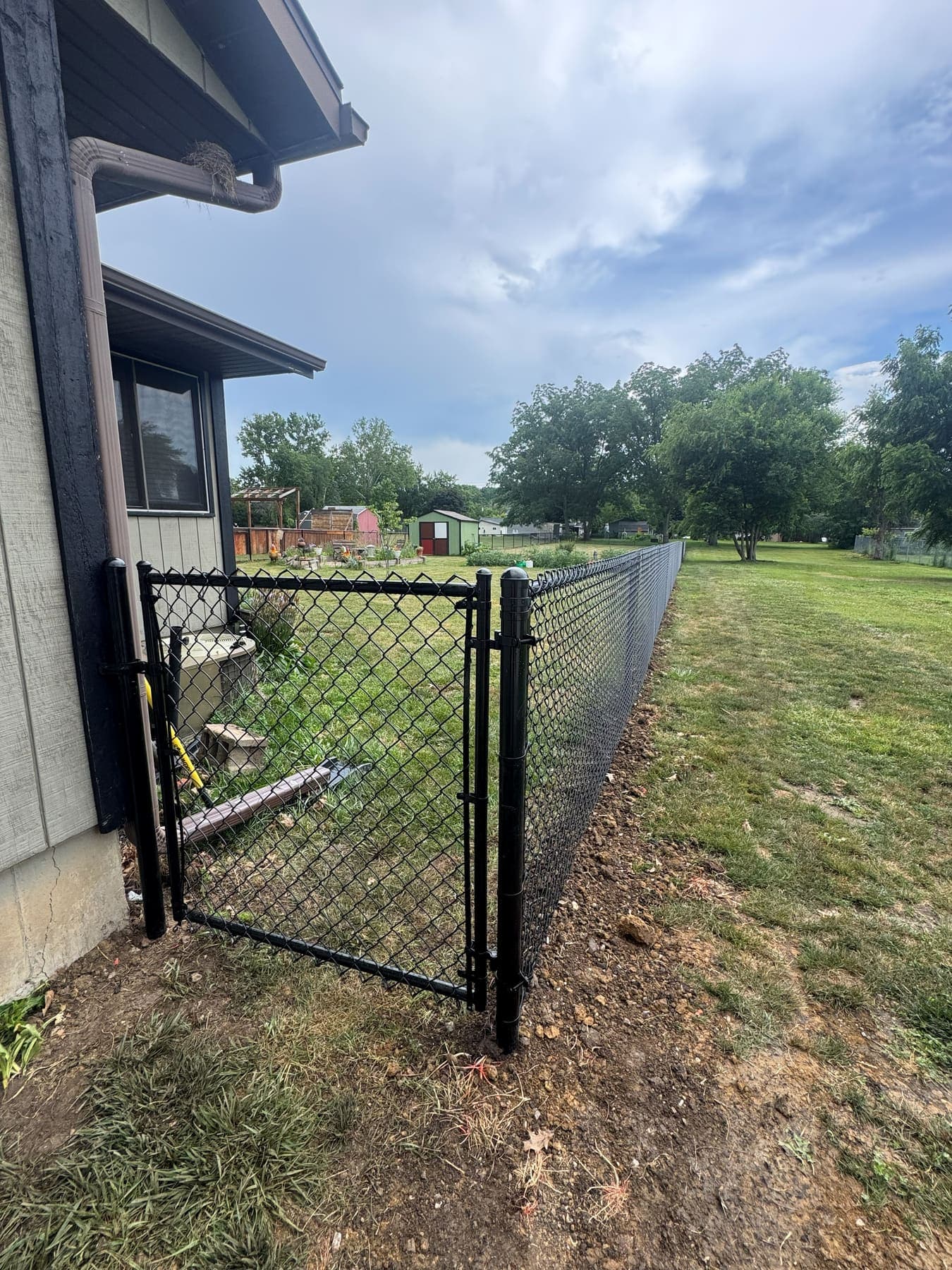 Black chain-link walk gate hung against a house, with the run carrying on into the yard behind it