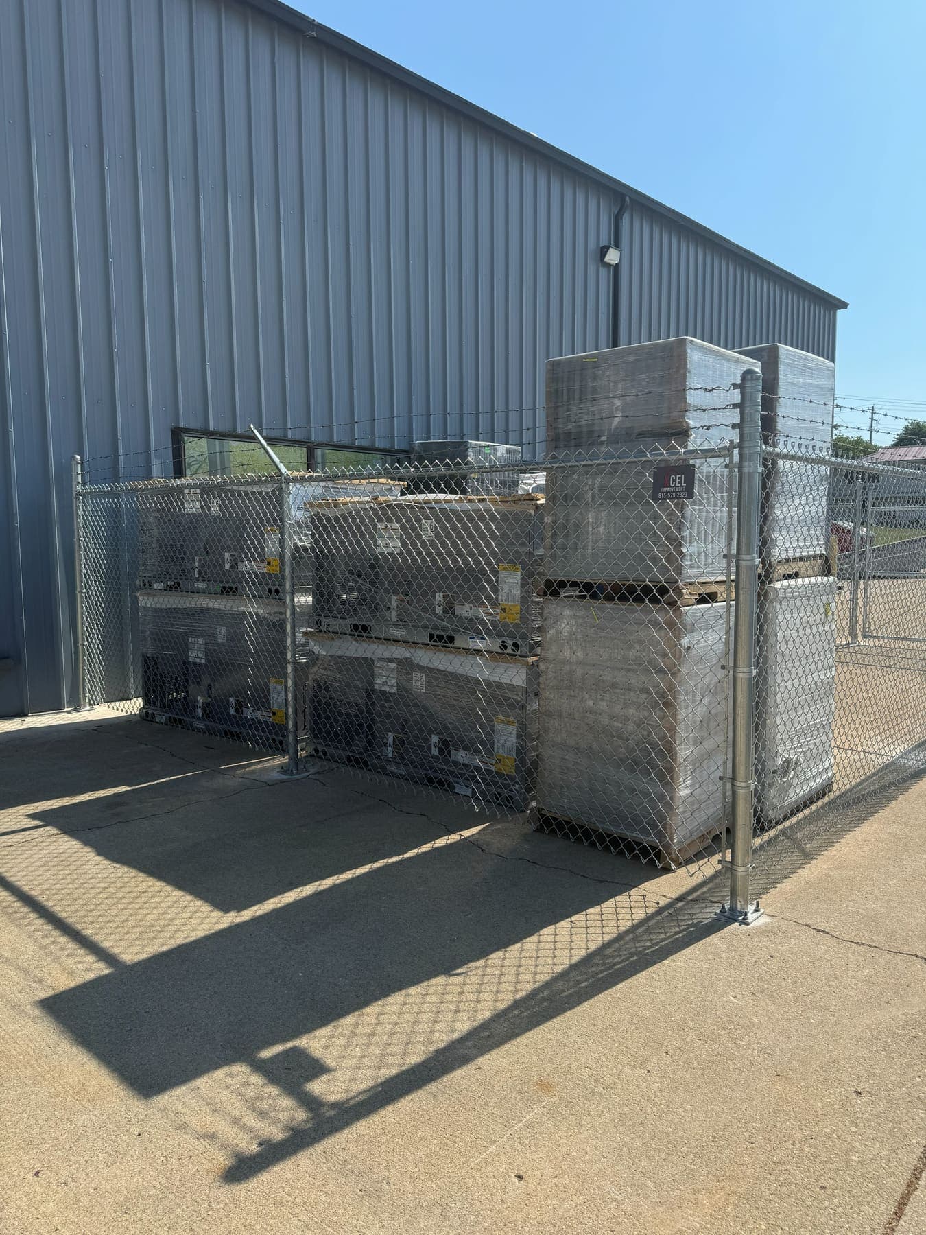 Galvanized chain-link security fence with three strands of barbed wire angled out along the top rail, enclosing a commercial storage yard of shrink-wrapped pallets beside a steel building, installed by Xcel Outdoor Living