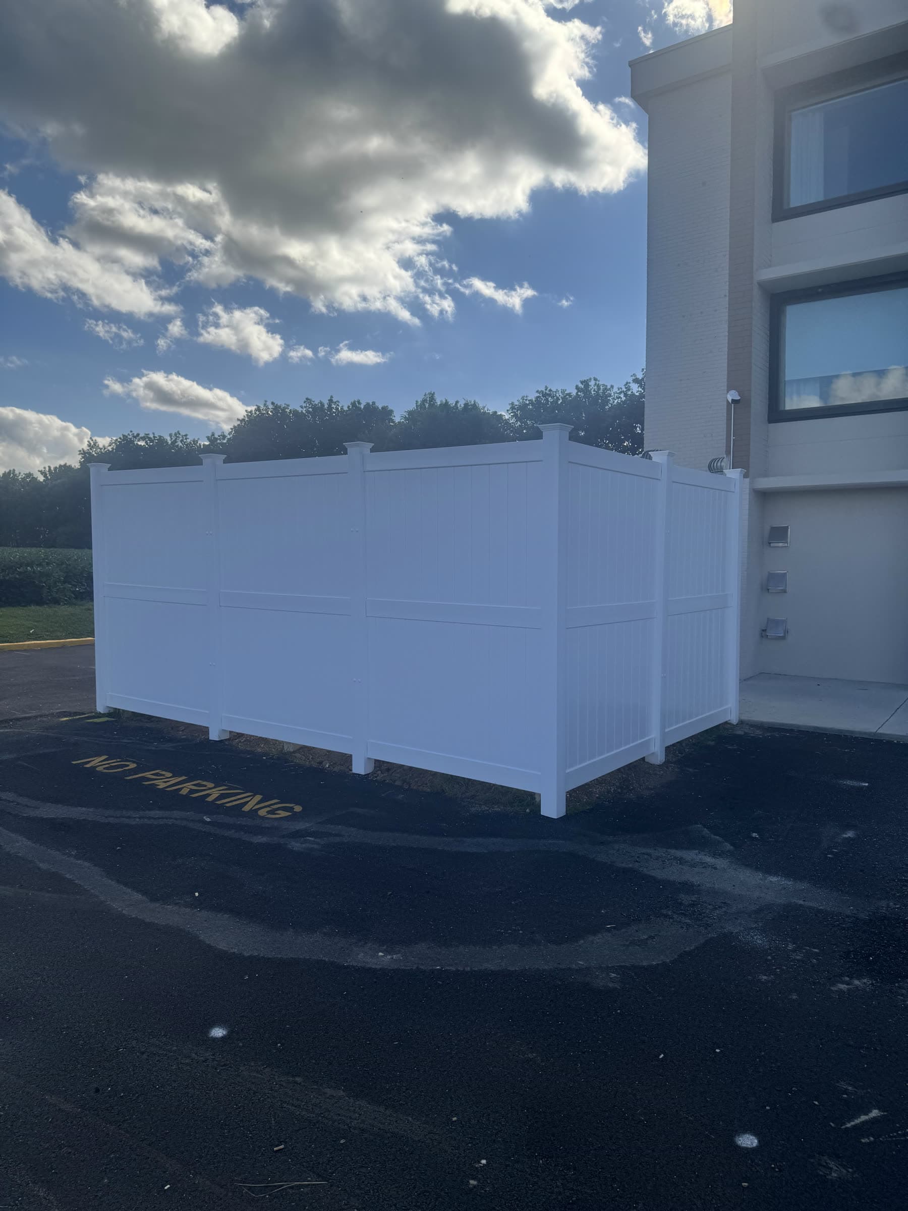 White vinyl privacy screen enclosure of three panel bays and a return, built on the asphalt at the corner of a brick commercial building by Xcel Outdoor Living