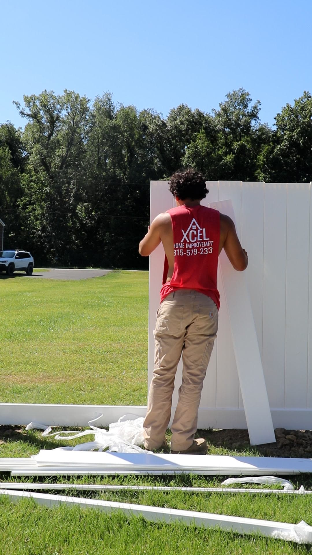 An Xcel crew member setting a white vinyl privacy panel into a fence run, with rails and panels stacked on the grass